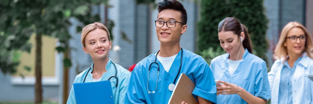 Group of medical students, including a man and women in scrubs, holding clipboards and walking outdoors with a confident stride.