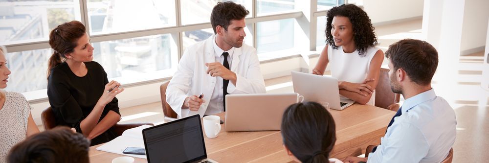 Five business professionals of diverse backgrounds sitting around a meeting table, engaged in a discussion with laptops and documents in front of them.
