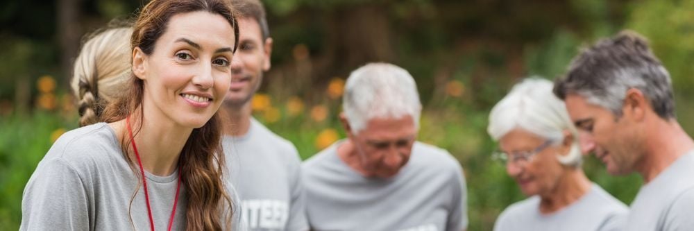 A diverse group of volunteers, including a young woman at the forefront, wearing gray t-shirts and lanyards, working together in a lush green park.