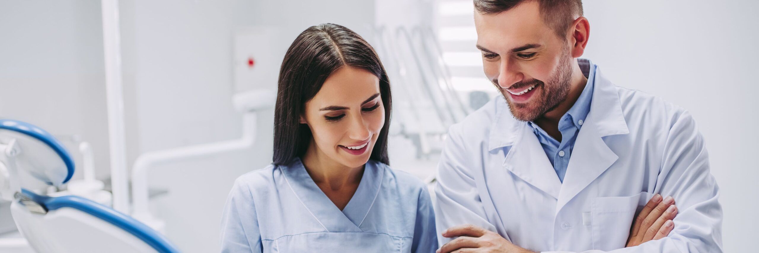 Two dental professionals, a man and a woman, smiling and looking at a digital tablet in a bright, modern dental clinic during their therapy classes.