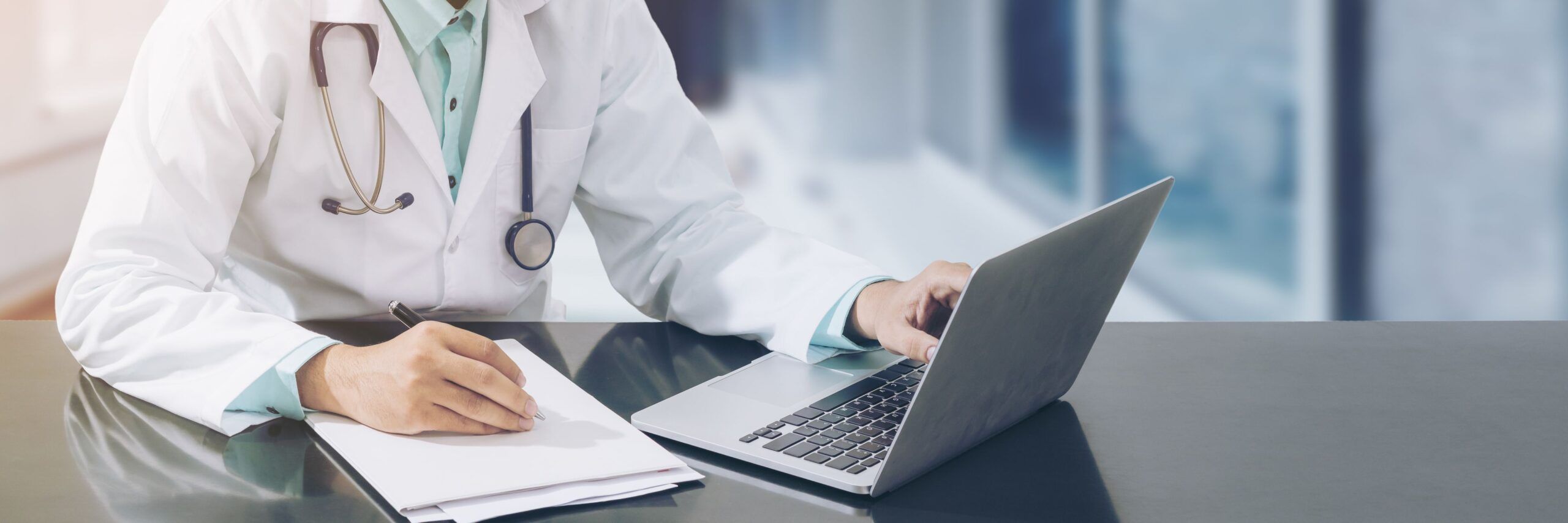 A doctor in a white coat with a stethoscope around his neck and a certification badge is using a laptop and writing notes at a desk in a modern office.