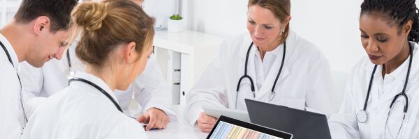 A diverse group of five healthcare students, wearing white lab coats and stethoscopes, engaged in a discussion around a table with laptops and student resources.