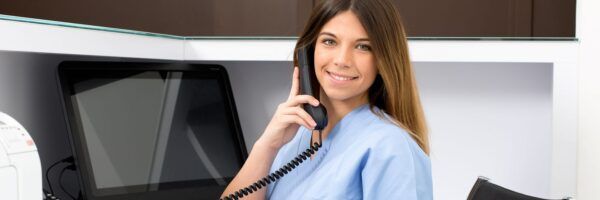 A smiling female medical student in blue scrubs holding a phone, standing in front of medical equipment in a clinic.