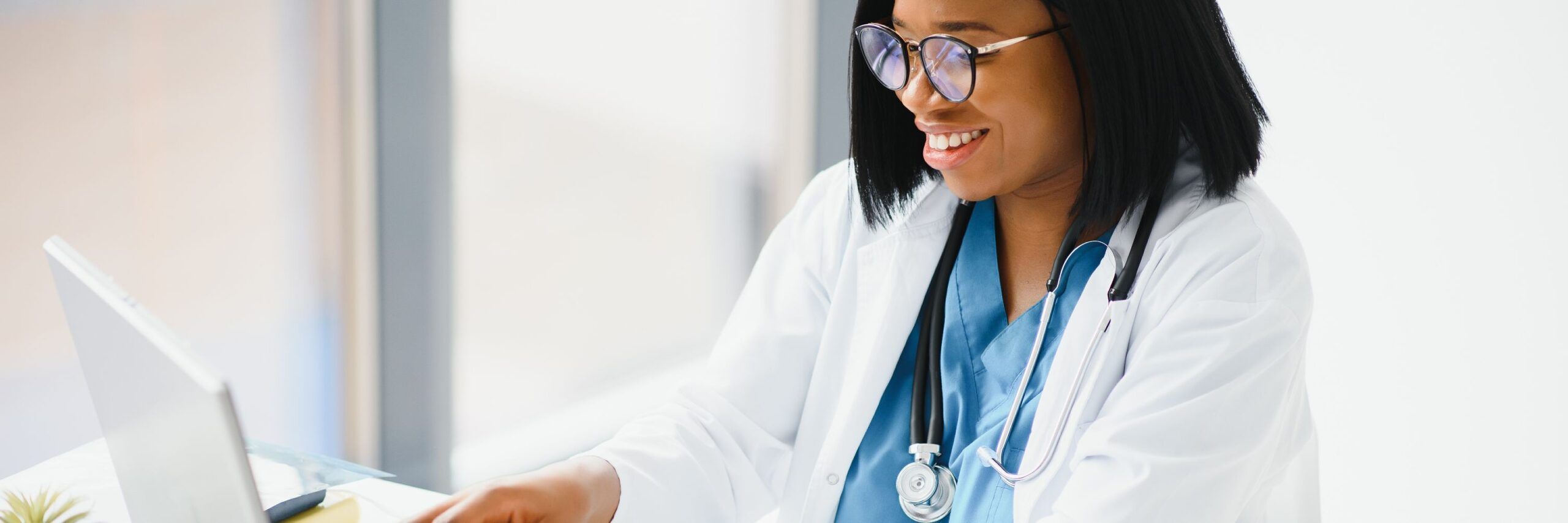 A smiling African American female doctor wearing glasses and a stethoscope uses student resources on a laptop in a brightly lit office.