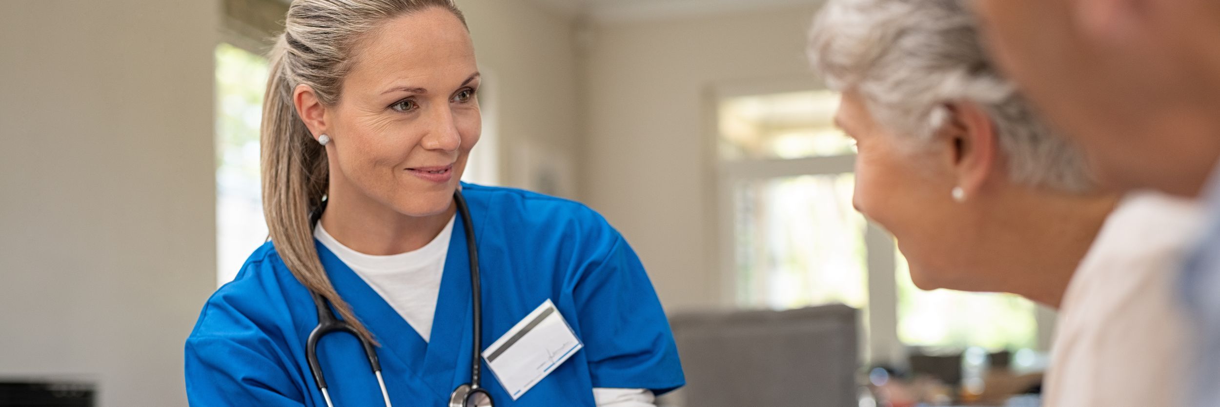 A healthcare professional with a Doctorate in Nursing Practice (DNP) in blue scrubs and a stethoscope engaging in a conversation with an elderly woman, both smiling in a bright indoor setting.