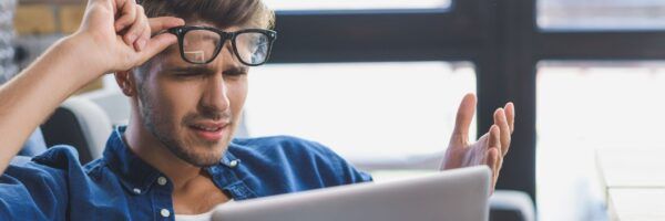 A perplexed man in a blue shirt wearing glasses, scratching his head while looking at a laptop screen with a confused expression as he plans his Hawaii vacation.
