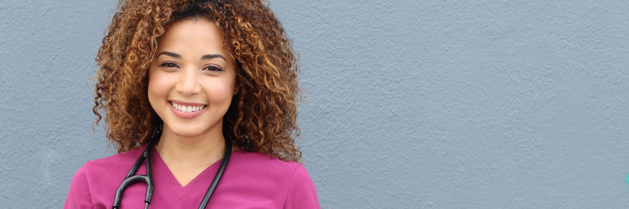 A smiling healthcare professional with curly hair wearing a pink scrub and a stethoscope around her neck, standing against a plain gray background.