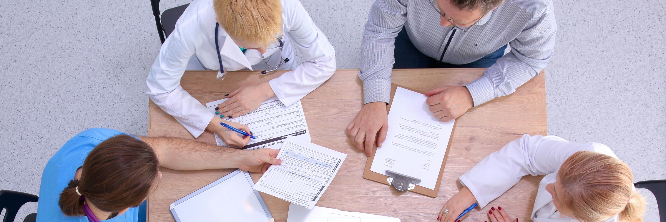 Top view of a group of five medical professionals discussing affordable documents at a rectangular table, using pens to write notes and review paperwork.