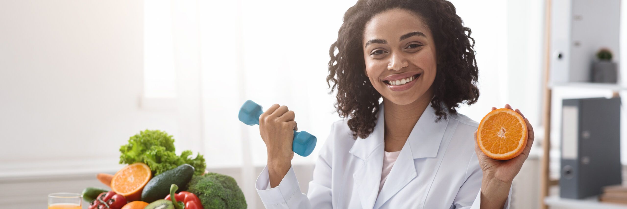 A smiling woman holding a dumbbell in one hand and half an orange in the other, seated at a table with fresh vegetables and fruits, promoting healthy living.