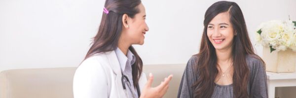 A female doctor in a white coat discussing with a smiling female patient in a grey top, seated in a well-lit, comfortable room with a bouquet on the side.