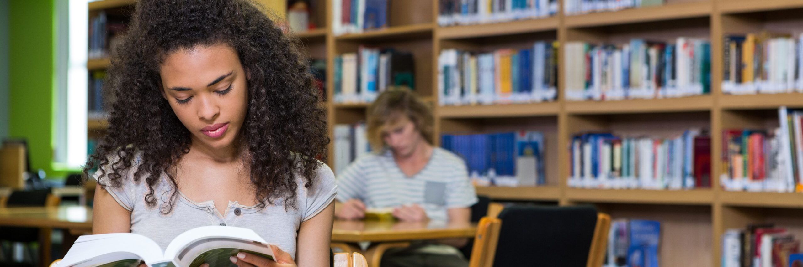 A young woman intently reads a book in a well-stocked library, with shelves of books and another person reading in the background.