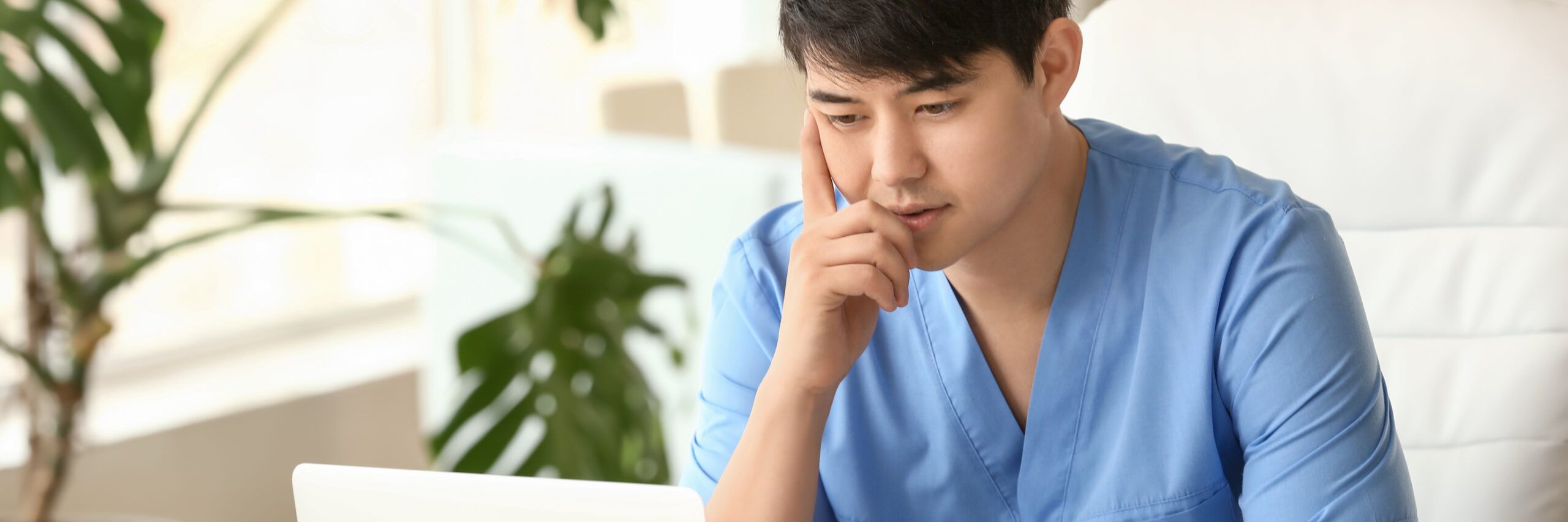 A thoughtful asian man in blue scrubs sits at a table with a laptop, touching his chin as he looks at the screen, with a white sofa and a plant in the background.