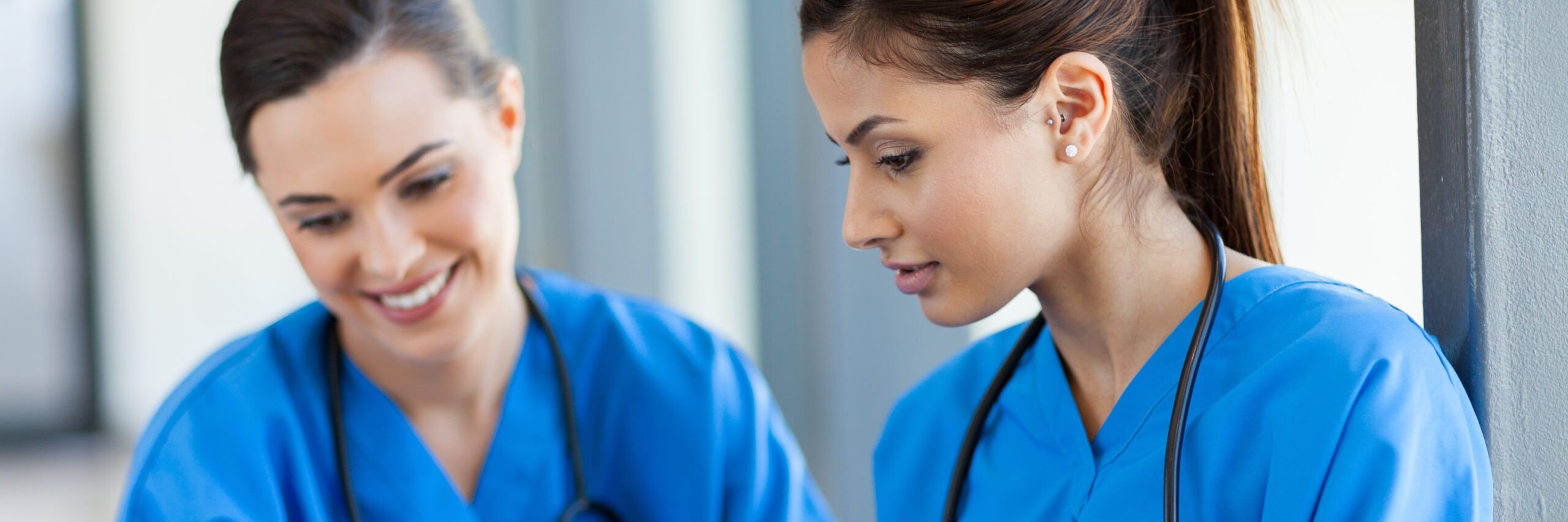 Two female healthcare professionals in blue scrubs, one smiling at the other as they engage in a discussion beside a window in a hospital corridor.