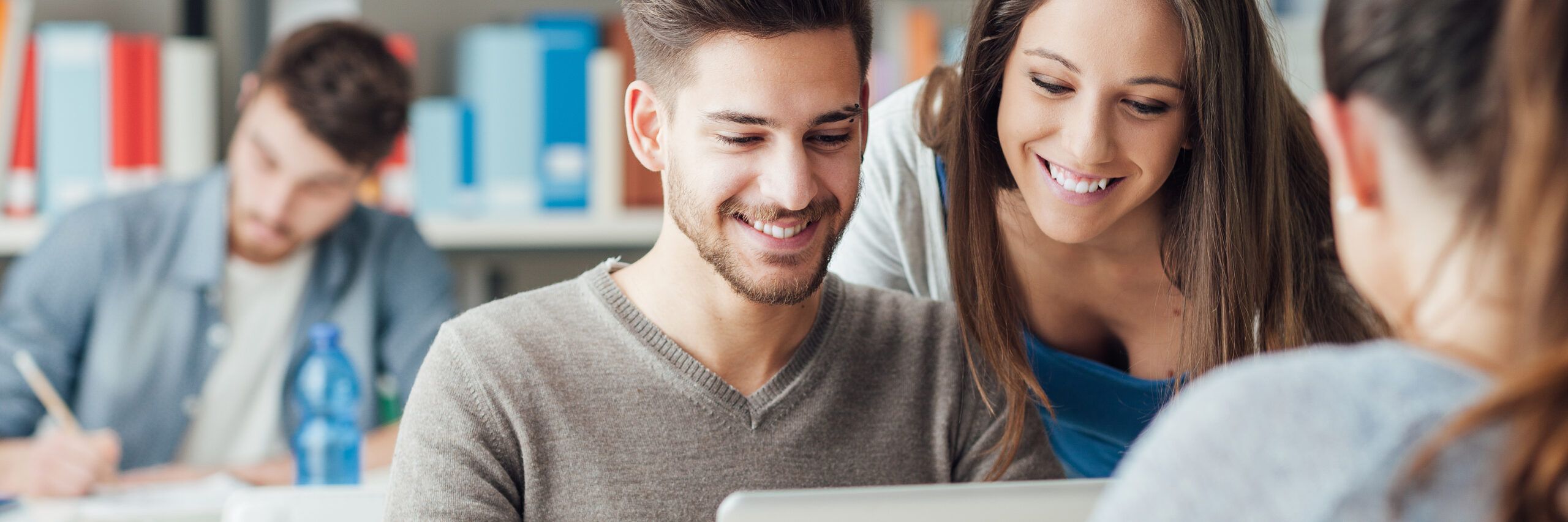 A group of young adults in a library, with a smiling man and woman looking at a tablet while another person studies in the background.