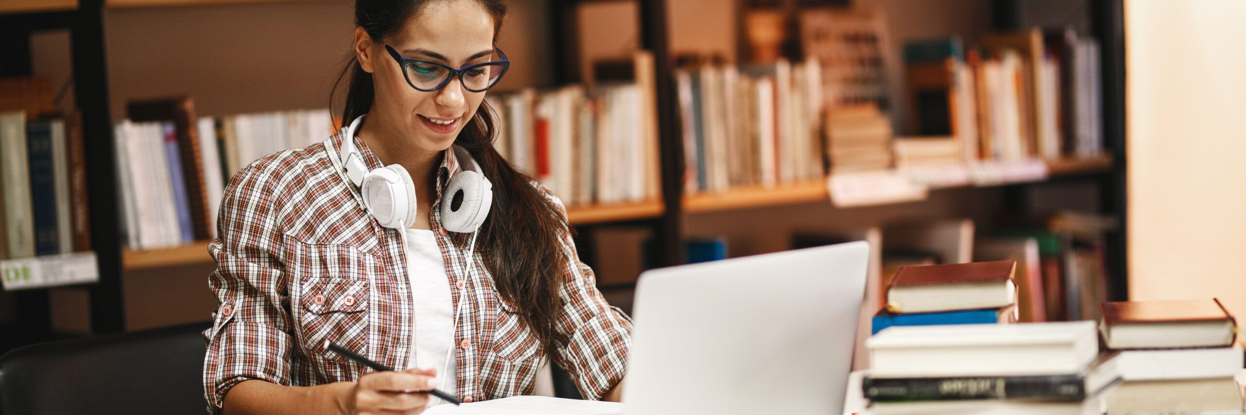 A smiling young woman wearing glasses and headphones around her neck studies intently at a laptop in a library, with books stacked around her.