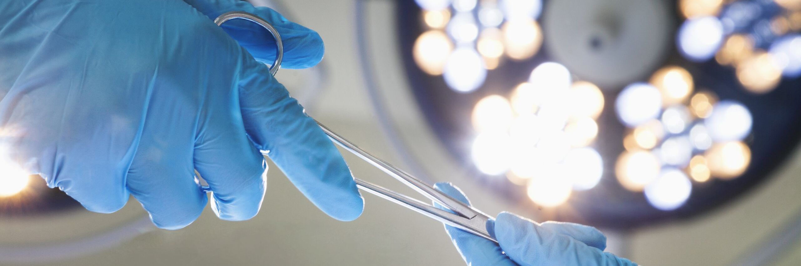 A close-up photo of a person wearing blue surgical gloves holding a surgical instrument, with out-of-focus surgical lights in the background.