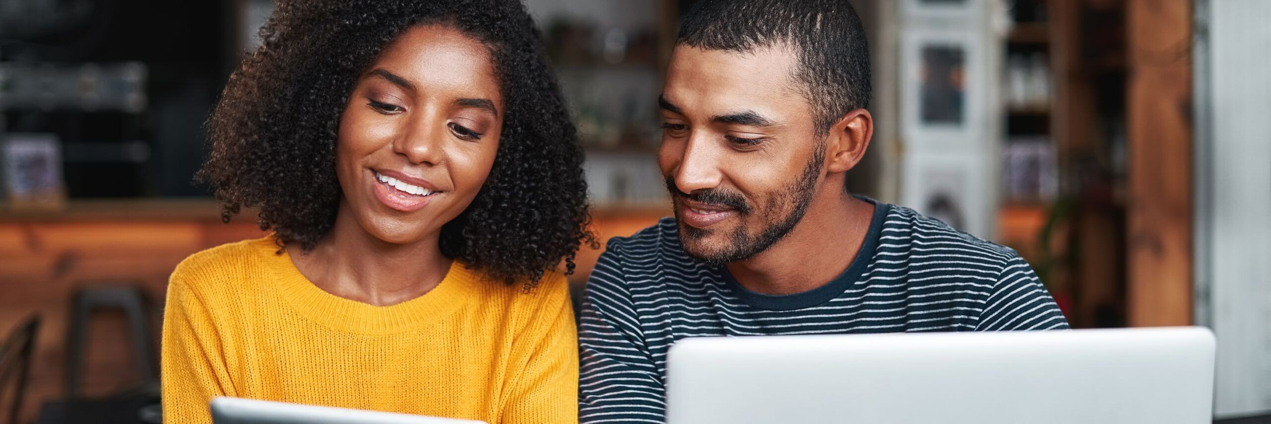 Two adults, a woman in a yellow sweater and a man in a striped shirt, smiling and looking at a laptop screen together in a cozy, well-lit setting.