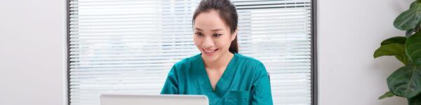 A female nurse practitioner in green scrubs smiles while using a laptop in a brightly lit office with blinds and a plant.