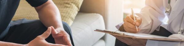 A person sits on a couch, attentively facing a medical professional in a white coat who is writing on a clipboard. The focus is on their hands and the clipboard, with sunlight streaming in from a window—symbolizing the journey from RN to PMHNP.