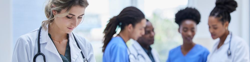 A group of four diverse female healthcare professionals, including doctors and nurse practitioners, engaged in discussion in a hospital corridor.