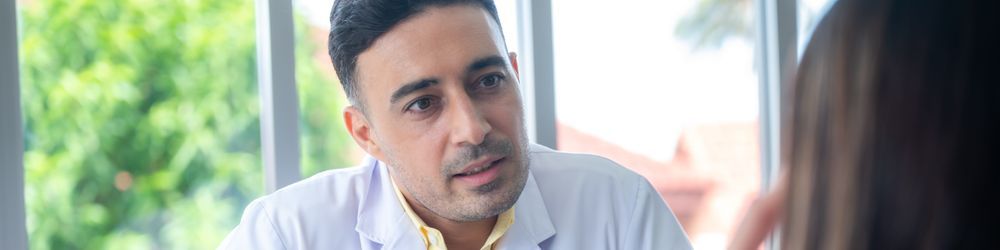 A doctor in a white coat is attentively listening to a patient during a consultation. The background features large windows with greenery and blurred outdoor scenery visible through them.