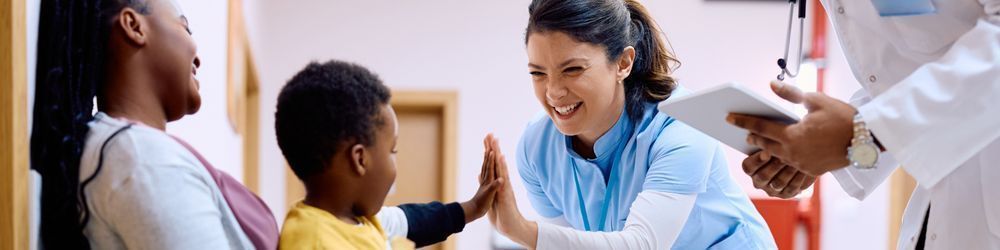 A smiling nurse in blue scrubs gives a high-five to a young boy sitting with his mother while a doctor with a tablet stands nearby. The interaction takes place in a medical setting, likely a clinic or hospital.
