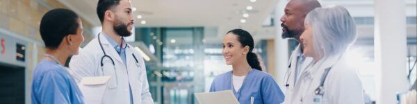 A diverse group of medical professionals, including a dedicated medical assistant, in scrubs and lab coats stand together, holding folders and engaging in a discussion about new opportunities in a bright hospital hallway.