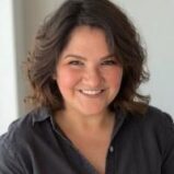 A woman with shoulder-length wavy brown hair and a warm smile, wearing a dark button-up shirt, poses in front of a light, neutral background.
