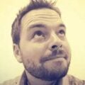 A man with short hair and a beard looks upward with a slight smile. The photo is in sepia tones, adding a warm, vintage effect.