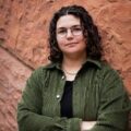 A person with curly dark hair, glasses, and a green corduroy jacket stands with arms crossed in front of a textured reddish-brown stone wall.