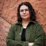A person with curly dark hair, glasses, and a green corduroy jacket stands with arms crossed in front of a textured reddish-brown stone wall.