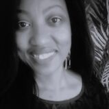 Black and white close-up photo of a woman smiling softly at the camera, with long dark hair and geometric earrings. Leafy patterned background partially visible on the right.