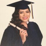 Young woman in a navy blue graduation cap and gown, smiling and holding a diploma scroll in her right hand. She has long wavy hair and is standing in front of a plain light background.