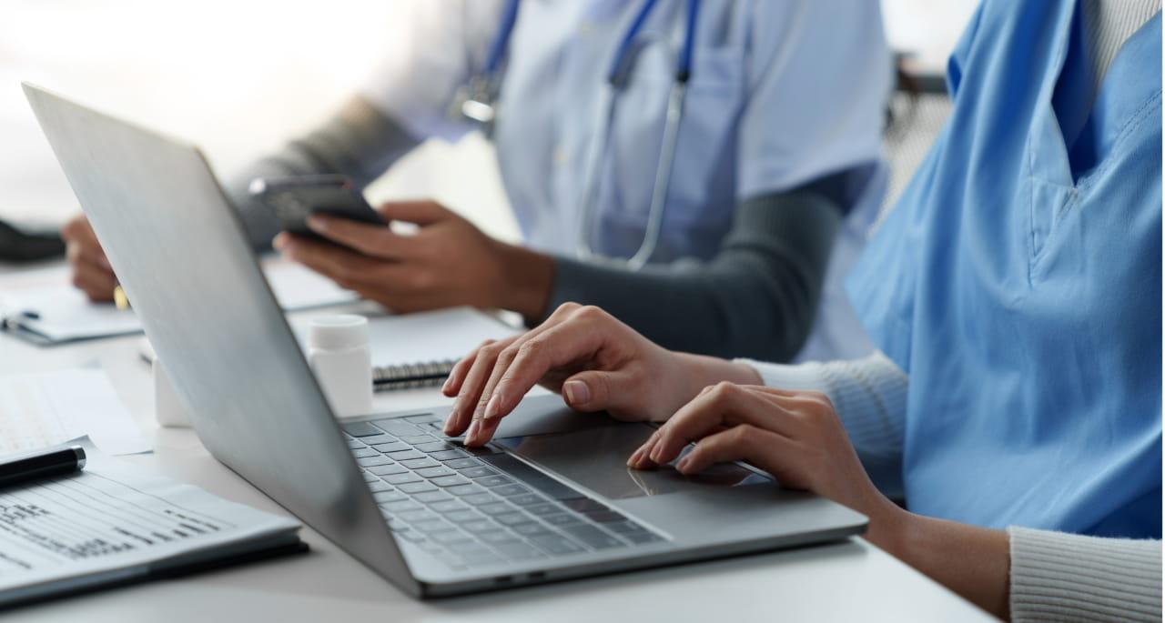 Two healthcare professionals in scrubs work at a desk; one types on a laptop, possibly reviewing NCLEX exam materials, while the other checks a smartphone. Medical documents and a stethoscope are visible on the table.