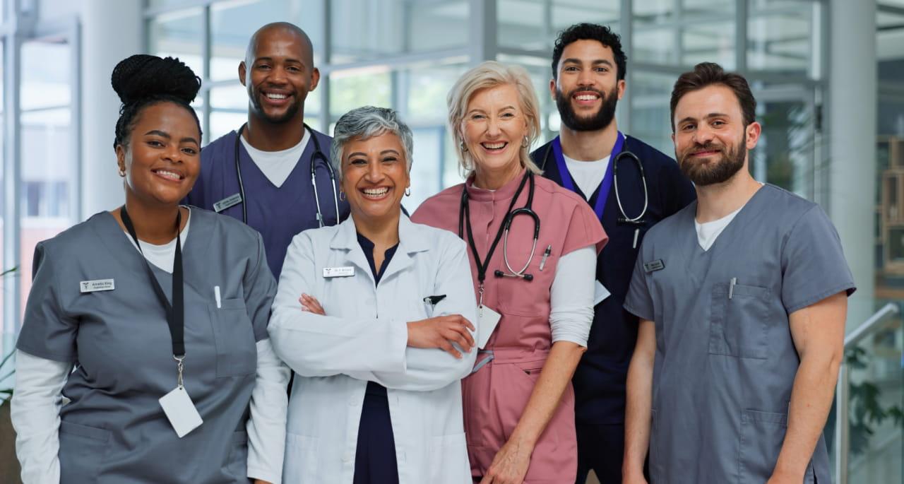 A diverse group of six smiling healthcare professionals, including doctors and nurses, stand together in a modern medical facility, wearing scrubs and lab coats with stethoscopes around their necks.