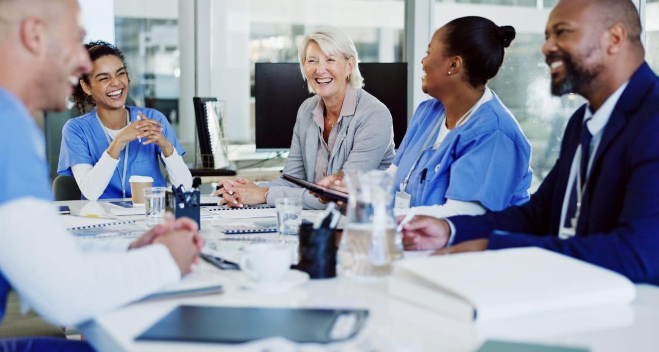 A group of healthcare professionals, including doctors and nurses, sit around a table in a brightly lit meeting room, smiling and having a discussion about nursing student writing. Notebooks, tablets, and drinks are on the table.