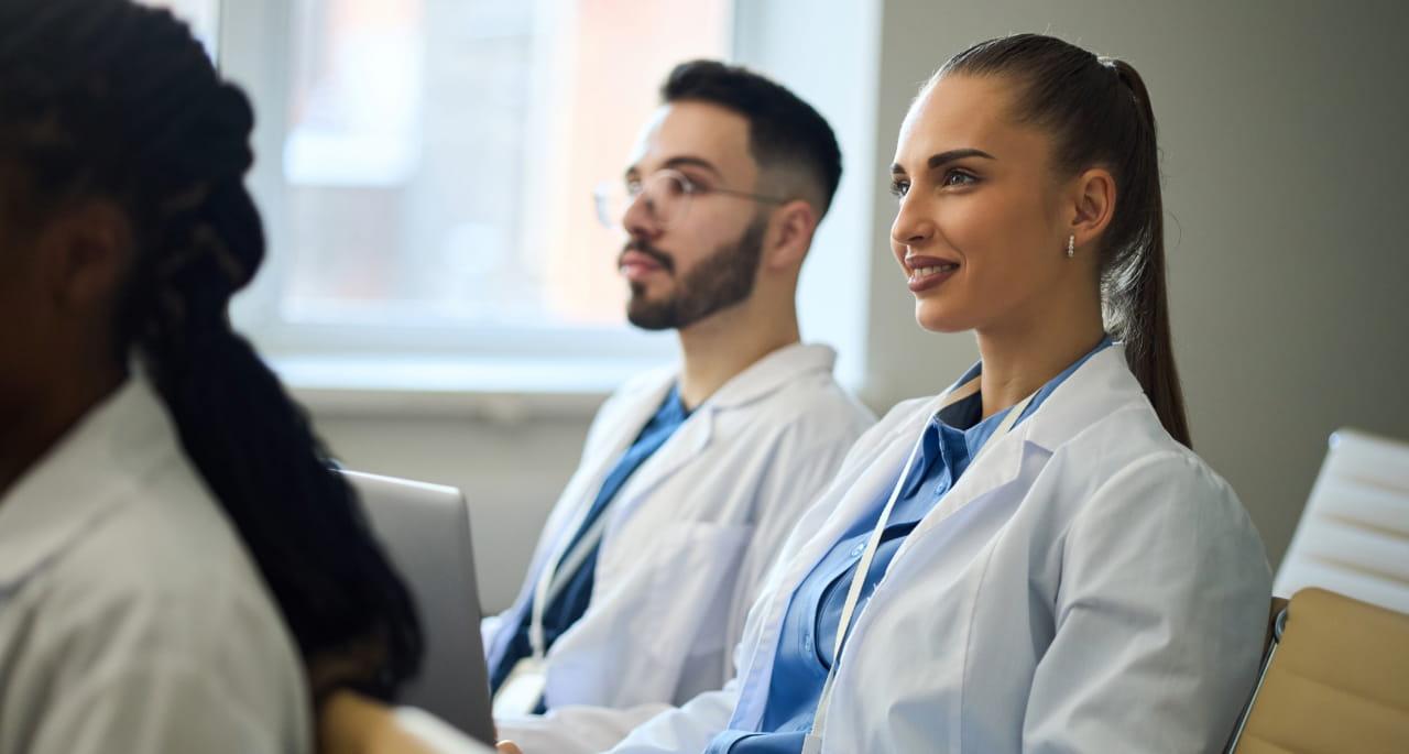Two medical professionals in white lab coats sit side by side in a bright room, attentively listening—perhaps discussing nursing school writing. One has long hair tied back, and both appear engaged, with a laptop visible in the background.