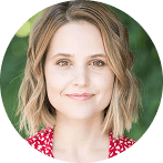 A woman with short, wavy blonde hair and light skin is smiling slightly. She is wearing a red top with a white pattern, and there is an unusual, blurred green background behind her.