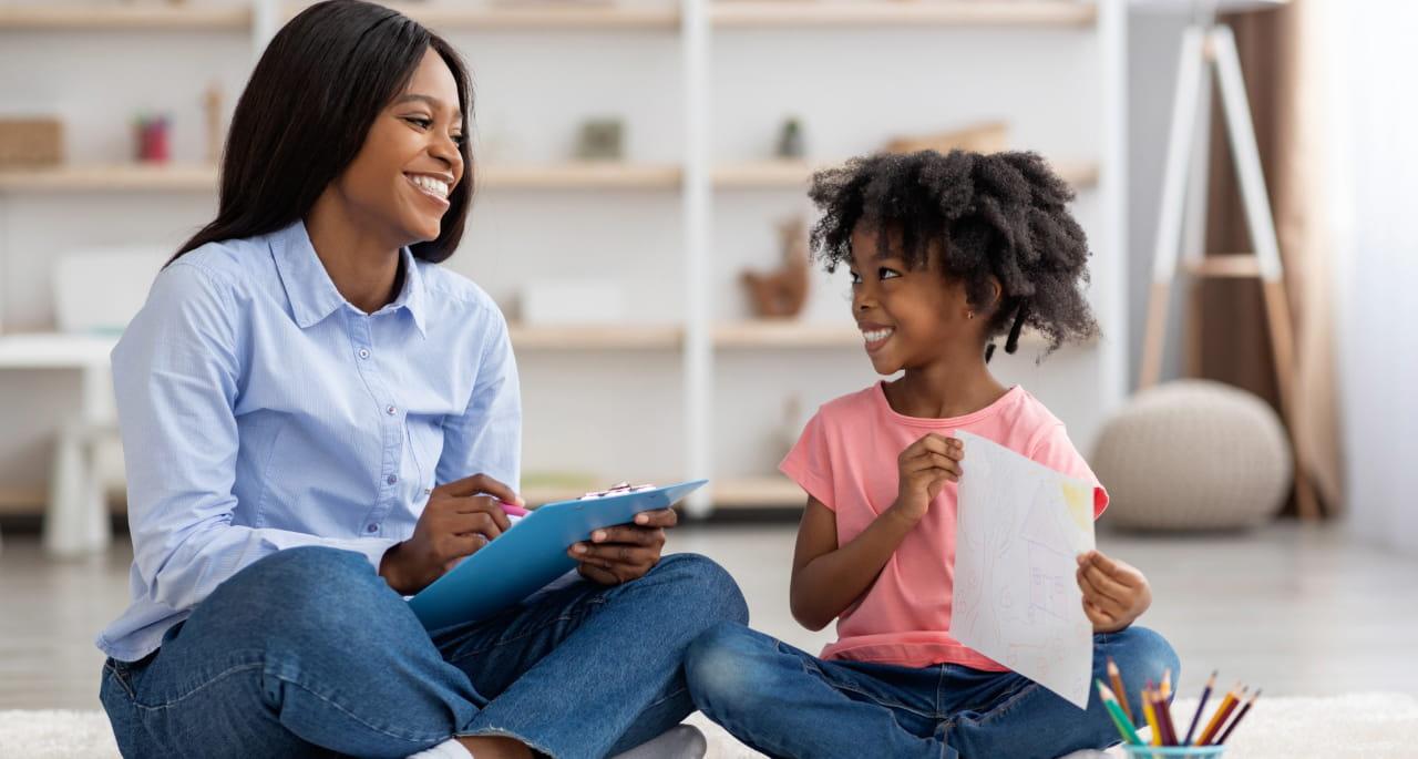 A woman and a young girl sit on the floor, smiling at each other. The woman holds a clipboard and pen, while the girl shows her an unusual drawing. Colored pencils and papers are scattered nearby.