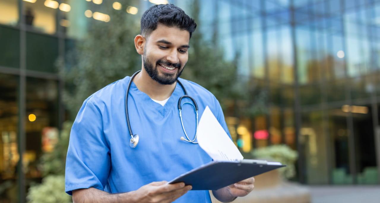 A smiling healthcare professional in blue scrubs with a stethoscope around his neck reviews nursing school writing on a clipboard outdoors near a modern glass building.