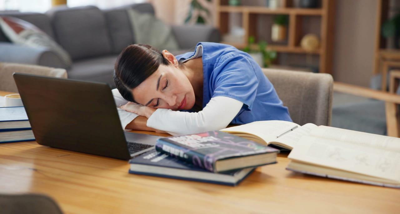 A woman in scrubs sleeps with her head on a book at a table cluttered with open textbooks and a laptop, suggesting fatigue from studying or working.