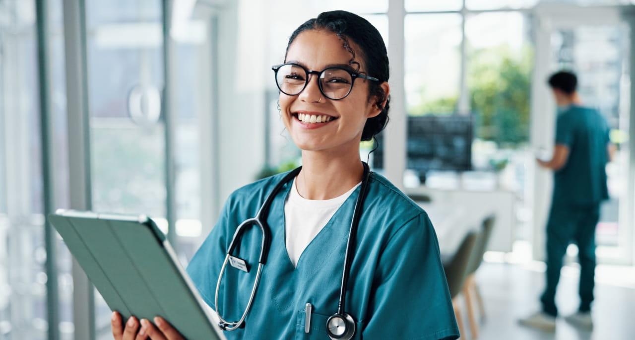 Smiling nurse in scrubs and glasses holds a digital tablet with a stethoscope around their neck, standing in a bright medical facility—highlighting the vital role of nurses and the impact of student loan forgiveness on healthcare professionals.