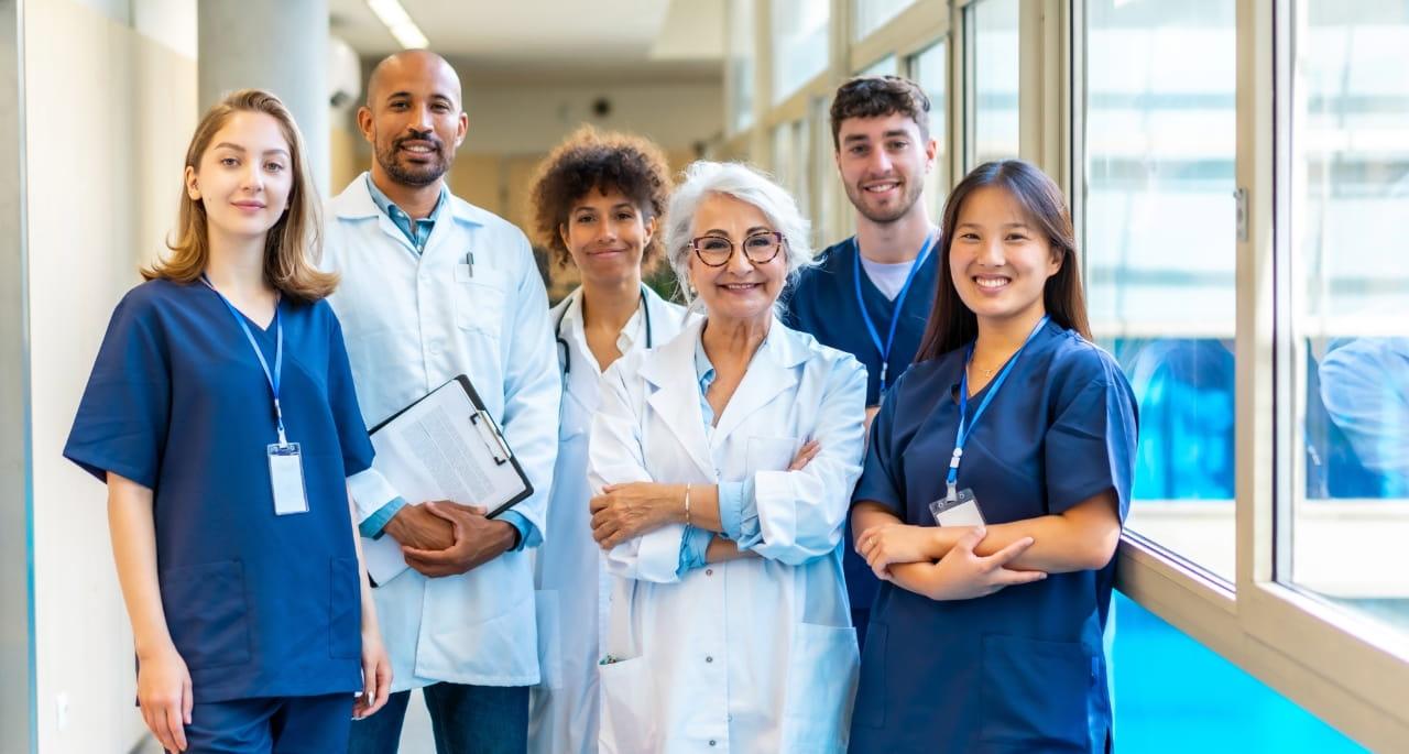 A group of six diverse healthcare professionals, including a nursing student writing notes, stand together smiling in a well-lit hospital hallway with large windows.