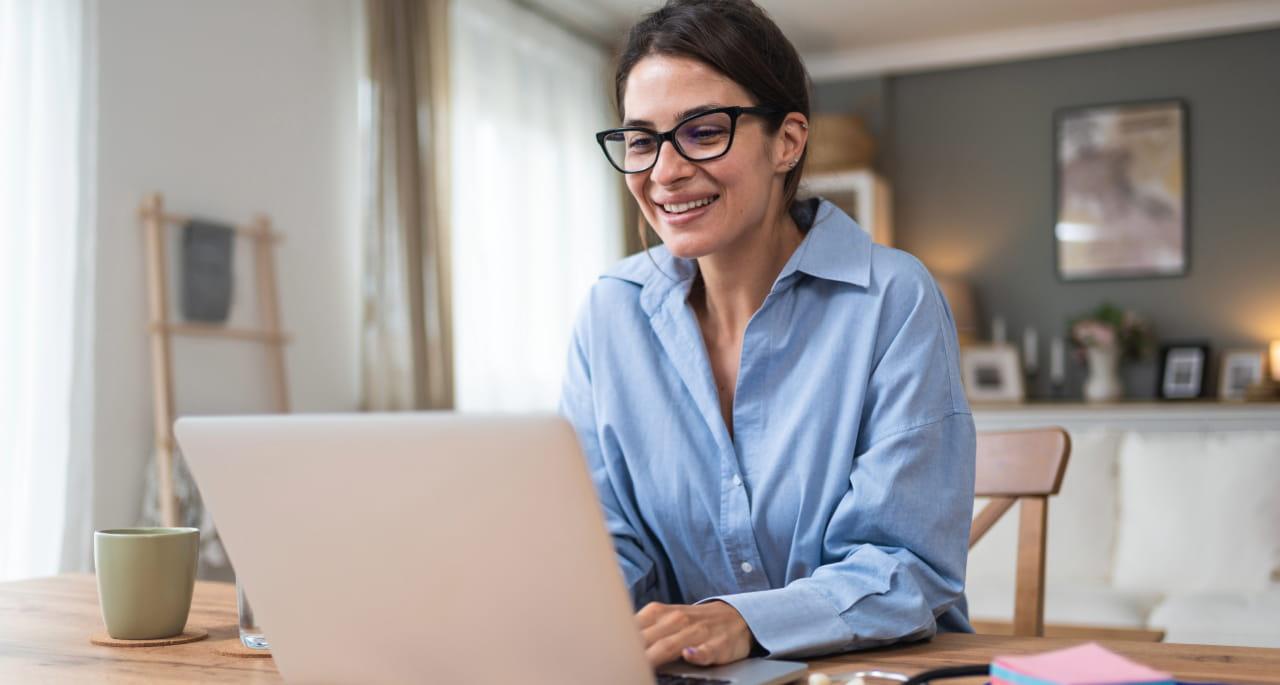 A woman wearing glasses and a blue shirt smiles while working on Nursing School Writing at her laptop, seated at a wooden table in a bright, cozy room with soft natural light.