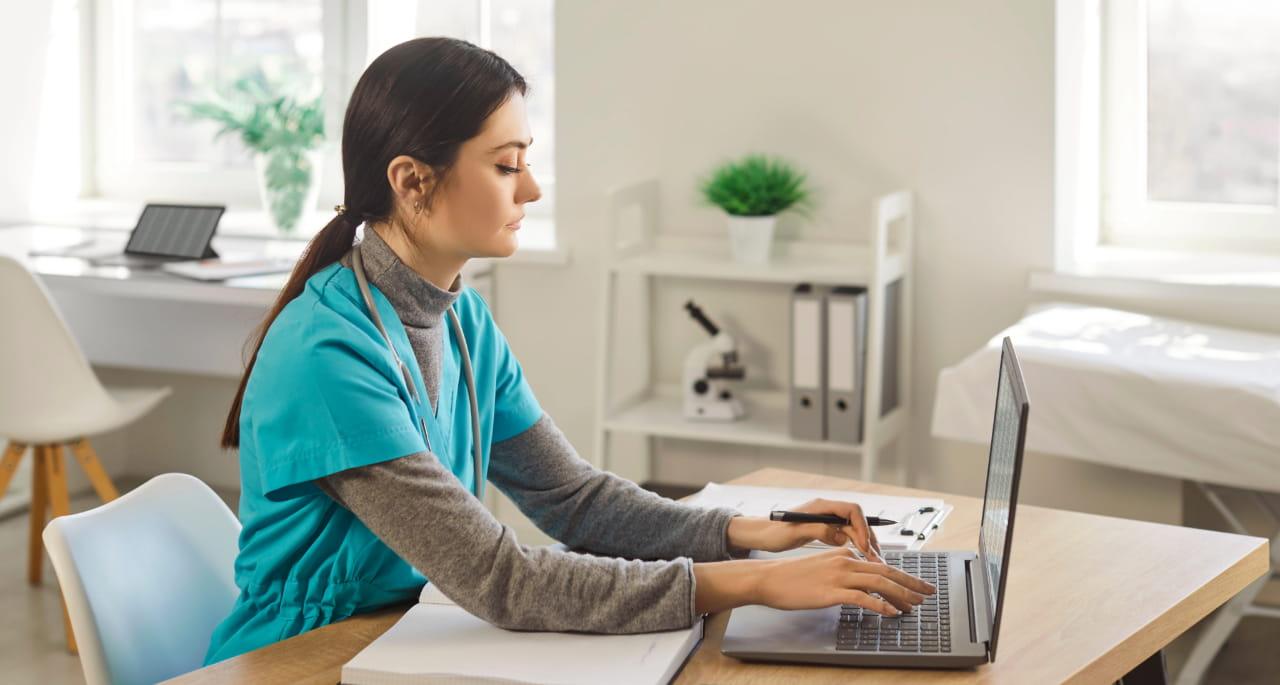 A healthcare worker in scrubs sits at a desk typing on a laptop in a bright, modern office with medical equipment, documents, and a plant in the background.