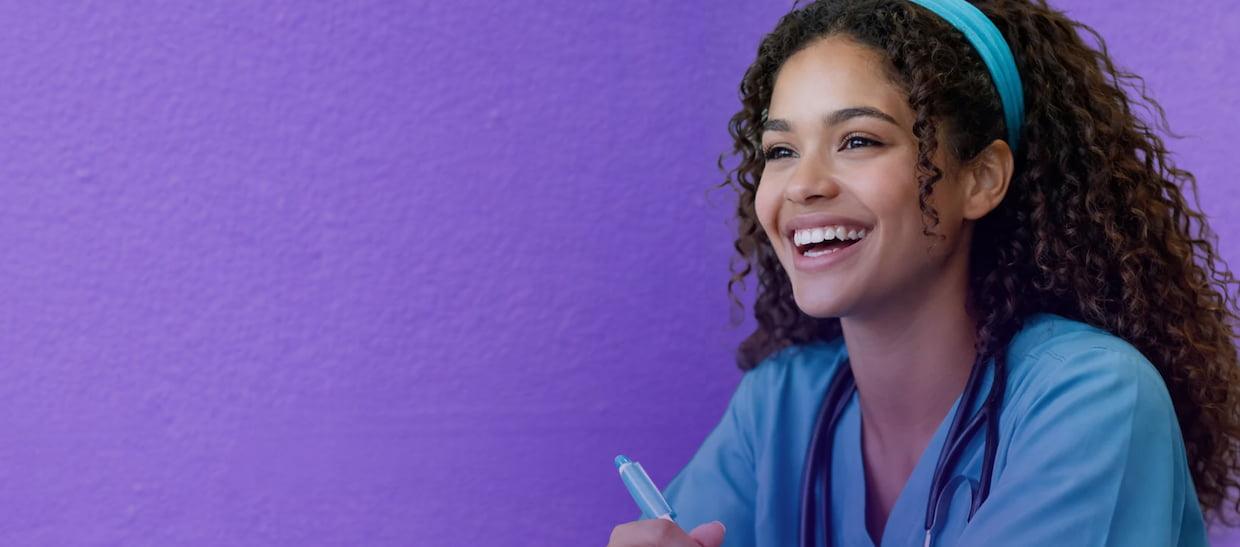 A young woman with curly hair, wearing medical scrubs and a stethoscope, smiles brightly while holding a pen. The background is a solid light purple wall.