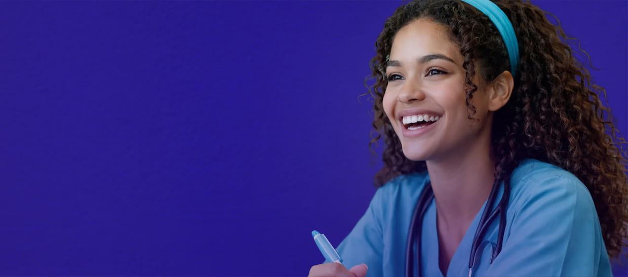 A smiling young woman in medical scrubs with a stethoscope around her neck holds a pen and sits against a solid purple background.